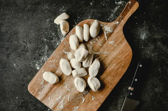 Raw dough pieces on a floured wooden cutting board next to a kitchen knife.